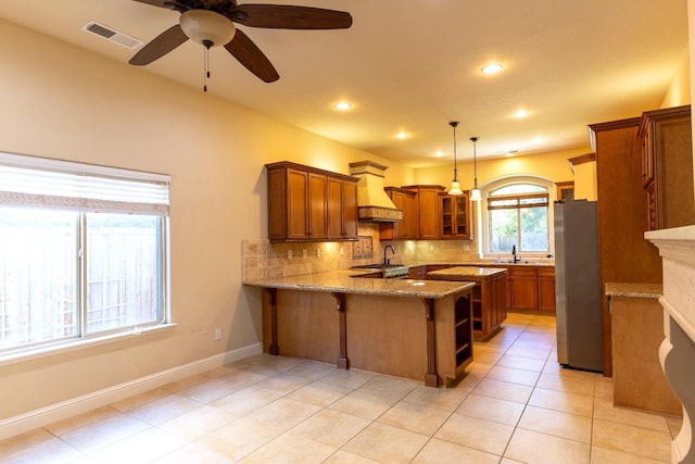 kitchen with custom range hood, tasteful backsplash, stainless steel refrigerator, light stone countertops, and kitchen peninsula