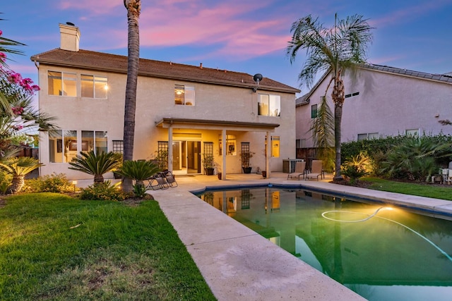 back of property at dusk with a yard, a chimney, stucco siding, a patio area, and an outdoor pool