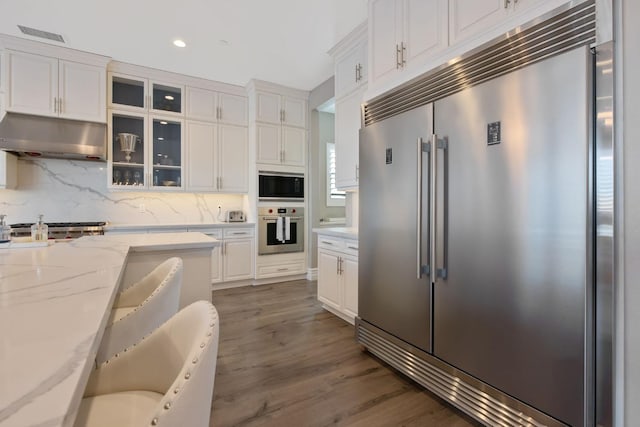 kitchen featuring built in appliances, decorative backsplash, hardwood / wood-style flooring, light stone counters, and white cabinets