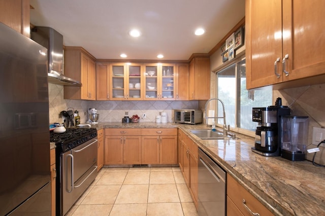 kitchen with glass insert cabinets, appliances with stainless steel finishes, a sink, light stone countertops, and wall chimney exhaust hood