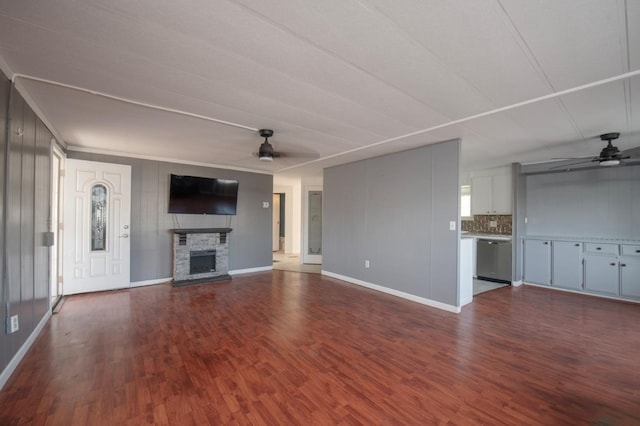 unfurnished living room featuring ornamental molding, a ceiling fan, dark wood finished floors, a stone fireplace, and baseboards