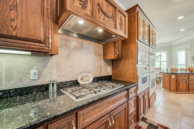 kitchen with dark stone counters, ornamental molding, stainless steel gas cooktop, light tile patterned floors, and custom range hood