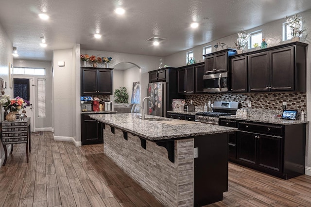 kitchen featuring an island with sink, light stone counters, appliances with stainless steel finishes, and hardwood / wood-style flooring