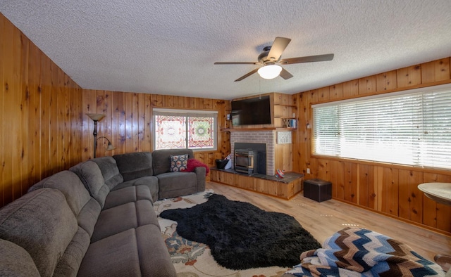 living room with wood walls, a textured ceiling, a wood stove, ceiling fan, and light hardwood / wood-style flooring