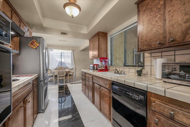 kitchen with backsplash, tile counters, sink, a tray ceiling, and black dishwasher