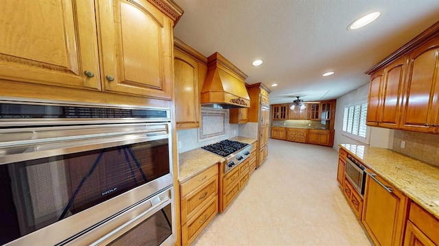 kitchen featuring light stone counters, custom exhaust hood, decorative backsplash, stainless steel appliances, and ceiling fan