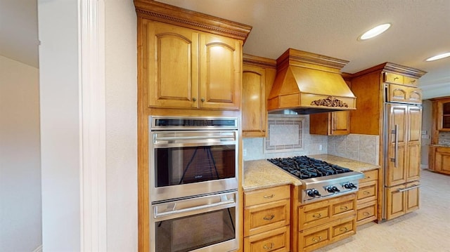 kitchen featuring backsplash, appliances with stainless steel finishes, light stone counters, and premium range hood