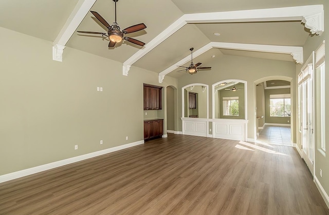 unfurnished living room with arched walkways, wood finished floors, a ceiling fan, baseboards, and beamed ceiling