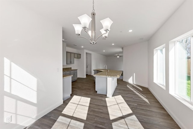 kitchen featuring dark wood-type flooring, a center island with sink, ceiling fan with notable chandelier, decorative light fixtures, and light stone counters