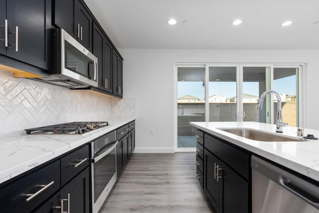 kitchen featuring stainless steel appliances, a sink, dark cabinetry, backsplash, and light stone counters