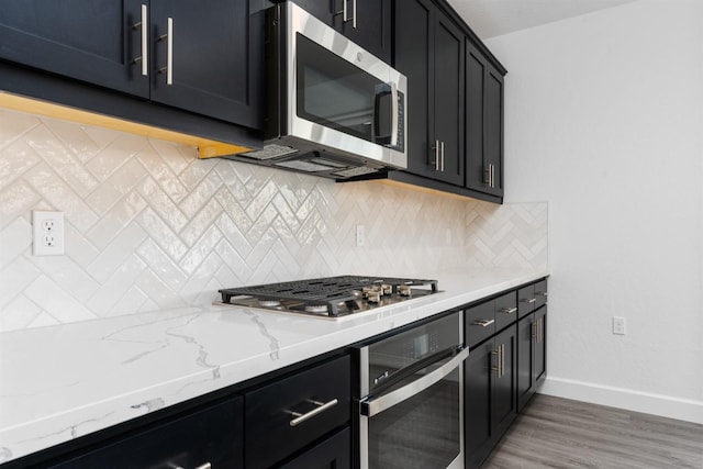 kitchen with backsplash, dark cabinetry, stainless steel appliances, baseboards, and light stone countertops