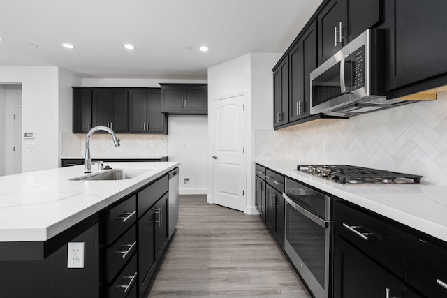 kitchen featuring a kitchen island with sink, a sink, light stone counters, dark cabinetry, and appliances with stainless steel finishes