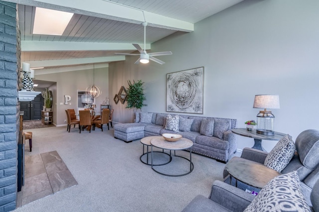 carpeted living room featuring a stone fireplace, vaulted ceiling with beams, and ceiling fan with notable chandelier