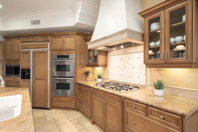 kitchen with custom range hood, decorative backsplash, appliances with stainless steel finishes, light stone counters, and crown molding