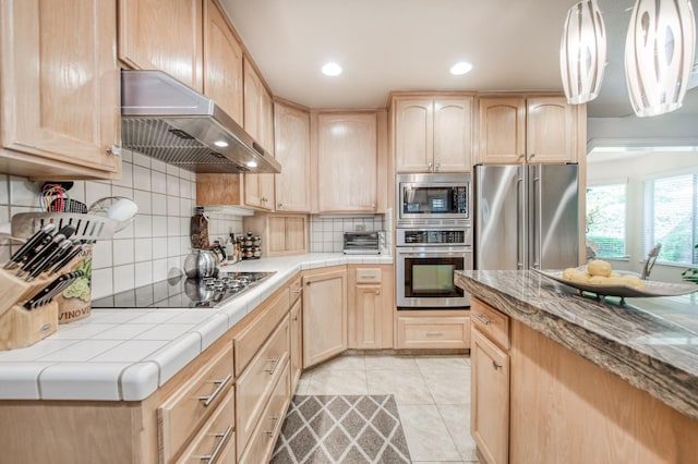 kitchen featuring tile counters, under cabinet range hood, decorative backsplash, stainless steel appliances, and light brown cabinetry