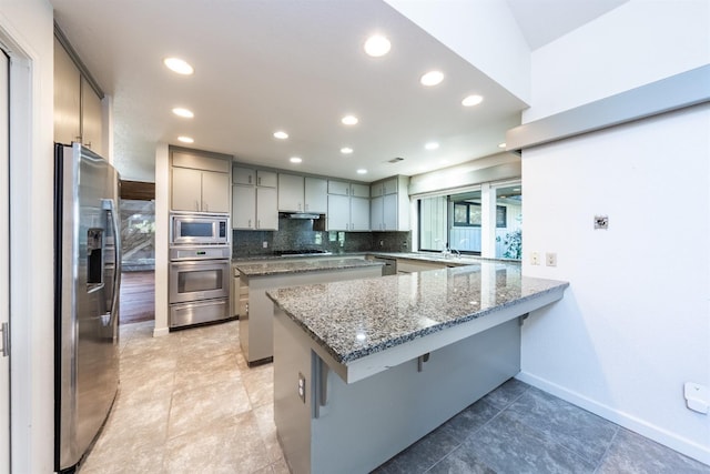 kitchen with gray cabinetry, kitchen peninsula, dark stone countertops, a kitchen bar, and appliances with stainless steel finishes