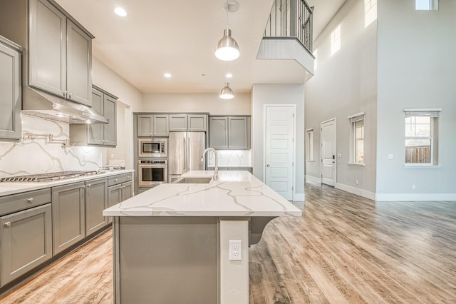 kitchen with gray cabinetry, under cabinet range hood, baseboards, a kitchen island with sink, and appliances with stainless steel finishes