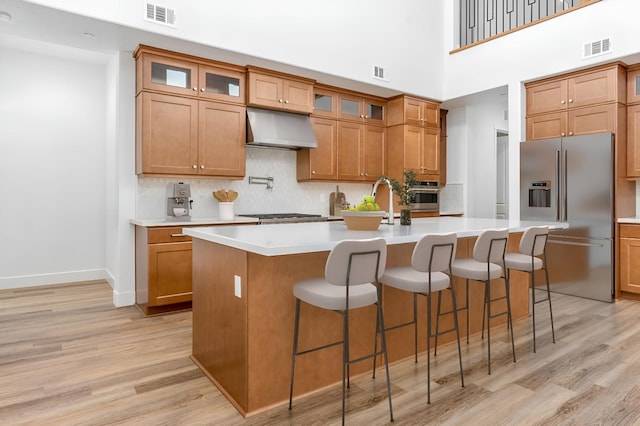 kitchen with stainless steel appliances, exhaust hood, light wood-type flooring, and an island with sink