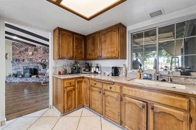kitchen with brown cabinetry, light stone countertops, a brick fireplace, a sink, and light tile patterned flooring