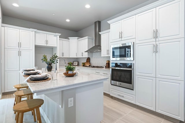 kitchen featuring wall chimney range hood, light stone countertops, appliances with stainless steel finishes, white cabinets, and a center island with sink