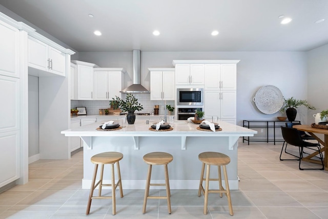 kitchen with wall chimney range hood, white cabinets, backsplash, appliances with stainless steel finishes, and a kitchen island with sink