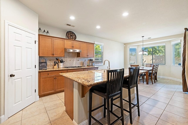 kitchen with a breakfast bar area, an island with sink, decorative light fixtures, light tile patterned floors, and tasteful backsplash