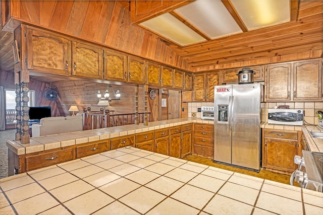 kitchen featuring tile counters, wooden walls, kitchen peninsula, stainless steel fridge with ice dispenser, and range