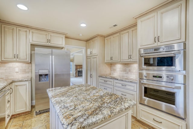 kitchen with a kitchen island, cream cabinets, backsplash, appliances with stainless steel finishes, and light stone counters