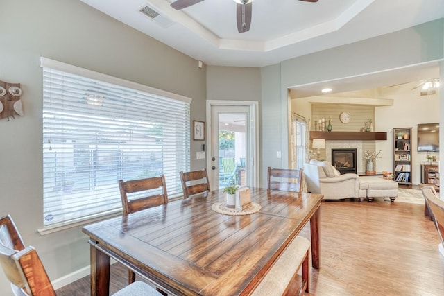 dining room featuring a stone fireplace, light hardwood / wood-style flooring, ceiling fan, and a tray ceiling