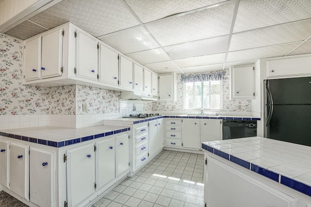 kitchen with wallpapered walls, white cabinets, black appliances, a drop ceiling, and a sink
