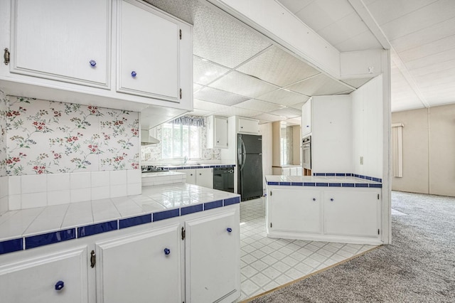 kitchen featuring white cabinets, black appliances, wallpapered walls, tile countertops, and light colored carpet