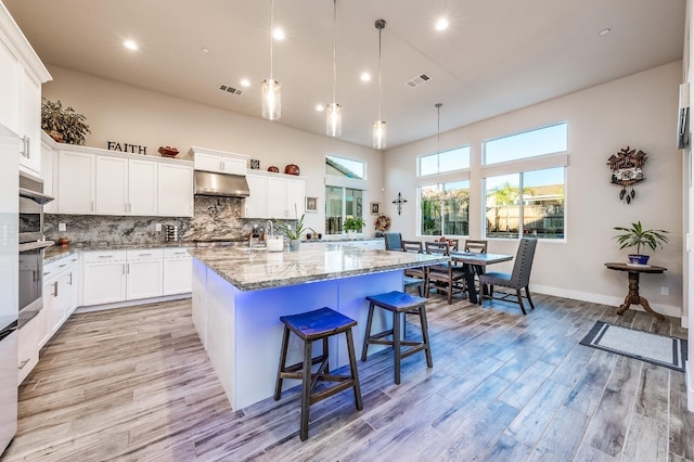 kitchen with under cabinet range hood, a kitchen island with sink, visible vents, and backsplash