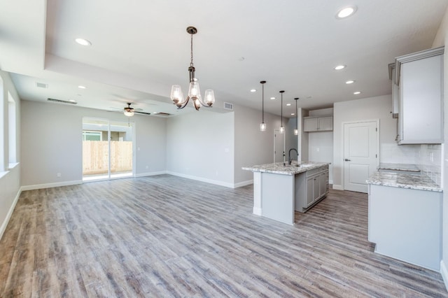 kitchen featuring decorative light fixtures, gray cabinetry, a kitchen island with sink, light hardwood / wood-style floors, and light stone countertops