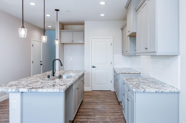 kitchen featuring sink, a kitchen island with sink, hanging light fixtures, light stone counters, and decorative backsplash