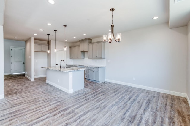 kitchen featuring gray cabinets, hanging light fixtures, a kitchen island with sink, light stone countertops, and light wood-type flooring