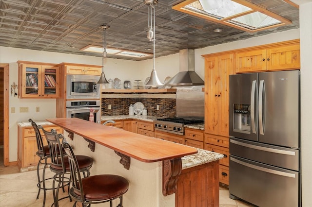 kitchen with a skylight, wall chimney exhaust hood, butcher block countertops, a center island with sink, and appliances with stainless steel finishes