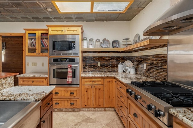 kitchen featuring exhaust hood, a skylight, decorative backsplash, light stone countertops, and stainless steel appliances