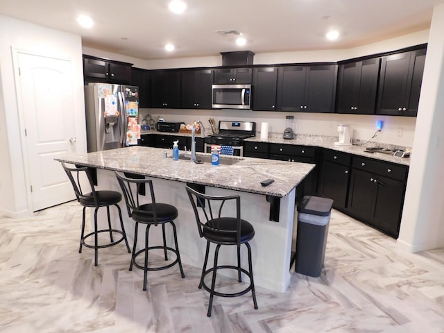 kitchen featuring a center island with sink, sink, light stone countertops, appliances with stainless steel finishes, and a breakfast bar area