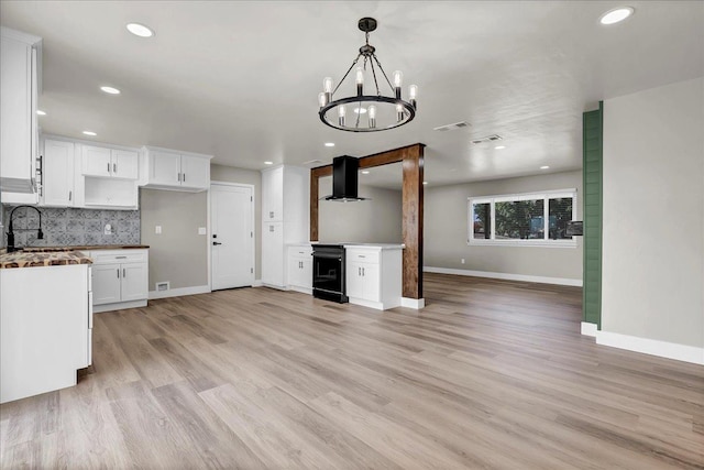 kitchen with light wood-type flooring, backsplash, wall chimney range hood, a notable chandelier, and white cabinets