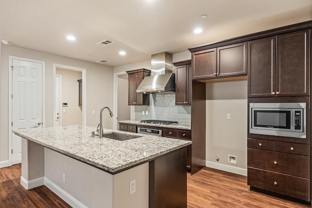 kitchen with wall chimney exhaust hood, sink, a kitchen island with sink, and appliances with stainless steel finishes