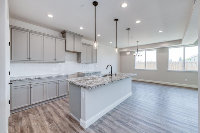 kitchen with light stone countertops, a center island with sink, gray cabinets, light hardwood / wood-style floors, and hanging light fixtures