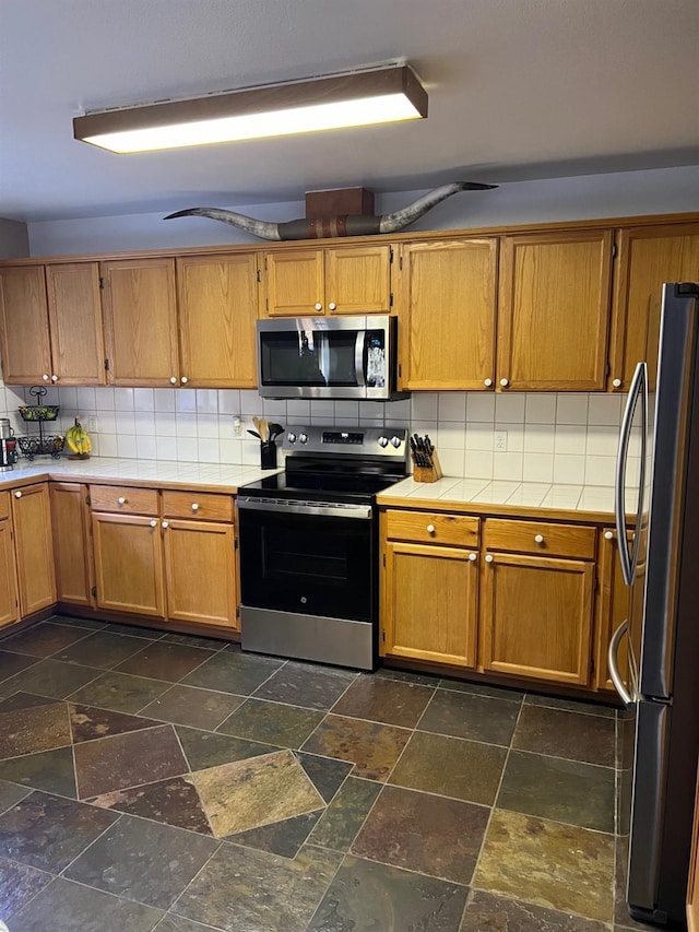 kitchen with stainless steel appliances, stone tile flooring, brown cabinetry, and backsplash