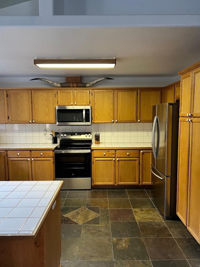 kitchen with appliances with stainless steel finishes, brown cabinetry, tile countertops, and tasteful backsplash