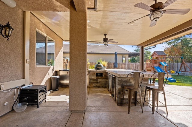 view of patio featuring a playground, outdoor dry bar, an outdoor kitchen, a ceiling fan, and fence