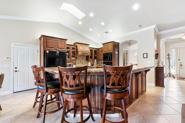 kitchen featuring black appliances, a kitchen bar, an island with sink, light stone counters, and lofted ceiling with skylight