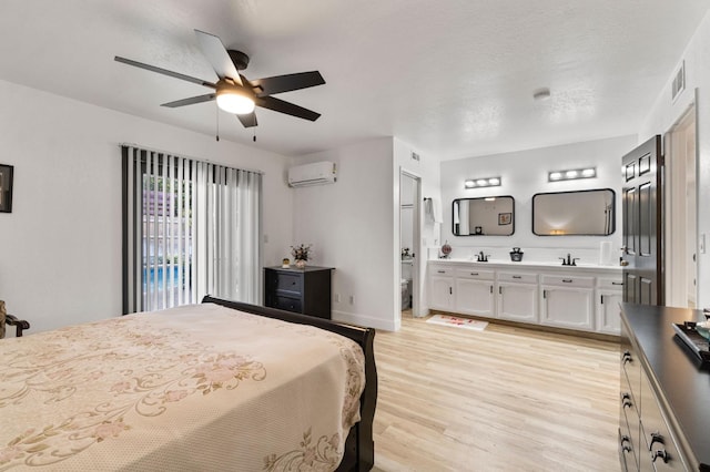 bedroom with ensuite bath, a textured ceiling, a wall unit AC, ceiling fan, and light hardwood / wood-style floors