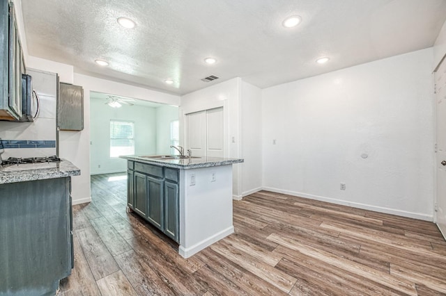 kitchen with light stone counters, ceiling fan, a kitchen island with sink, sink, and hardwood / wood-style floors