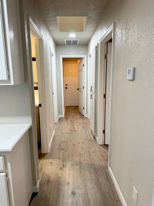 corridor with visible vents, a textured wall, light wood-style flooring, and baseboards