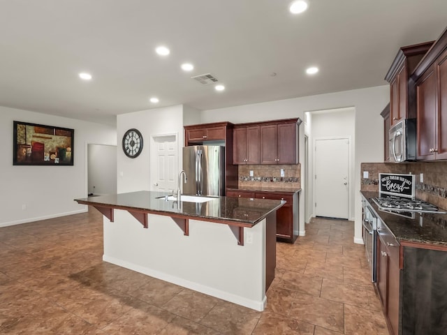 kitchen featuring stainless steel appliances, tasteful backsplash, dark stone countertops, an island with sink, and a breakfast bar area