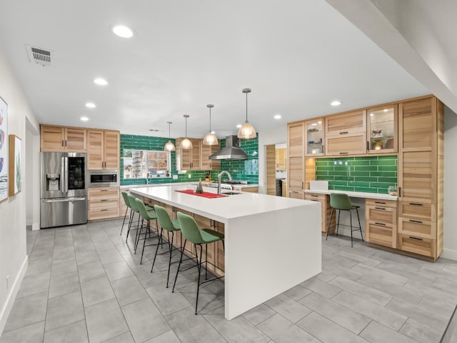 kitchen with backsplash, wall chimney range hood, an island with sink, appliances with stainless steel finishes, and a kitchen bar